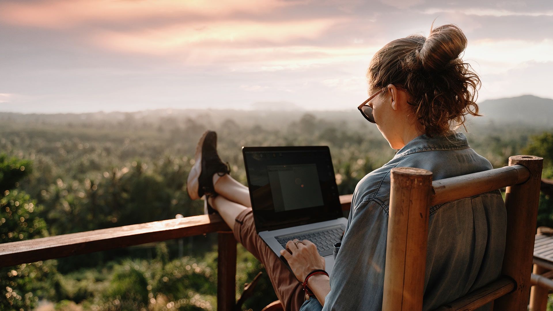 Eine Frau sitzt auf einer Veranda und arbeitet mit einem Laptop inmitten einer natuerlichen Umgebung.
